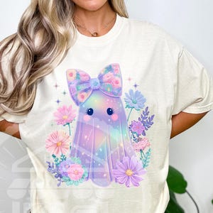 Pode incluir: Camiseta creme com um design de fantasma fofo. O fantasma &eacute; adornado com um la&ccedil;o floral e cercado por flores em tons pastel. O design tem um esquema de cores do arco-&iacute;ris, com tons de rosa, roxo e azul. A camisa &eacute; um estilo casual e confort&aacute;vel.