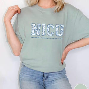 May include: A light green t-shirt with a floral print design that spells out "NICU" in large letters. The text below the letters reads "neonatal intensive care unit".