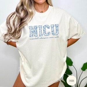 May include: A white t-shirt with a blue floral pattern spelling out "NICU" and the text "neonatal intensive care unit" below. The shirt is available in ivory.