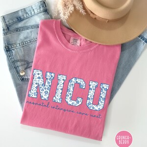 May include: Pink t-shirt with a blue and white floral print of the word "NICU" and the text "neonatal intensive care unit" in white.