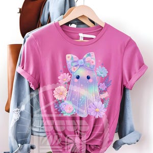 Pode incluir: Uma camiseta rosa com um desenho de fantasma de desenho animado. O fantasma tem um la&ccedil;o floral e &eacute; cercado por flores coloridas. O corpo do fantasma tem um efeito de gradiente de arco-&iacute;ris. A camiseta &eacute; de manga curta e gola redonda.