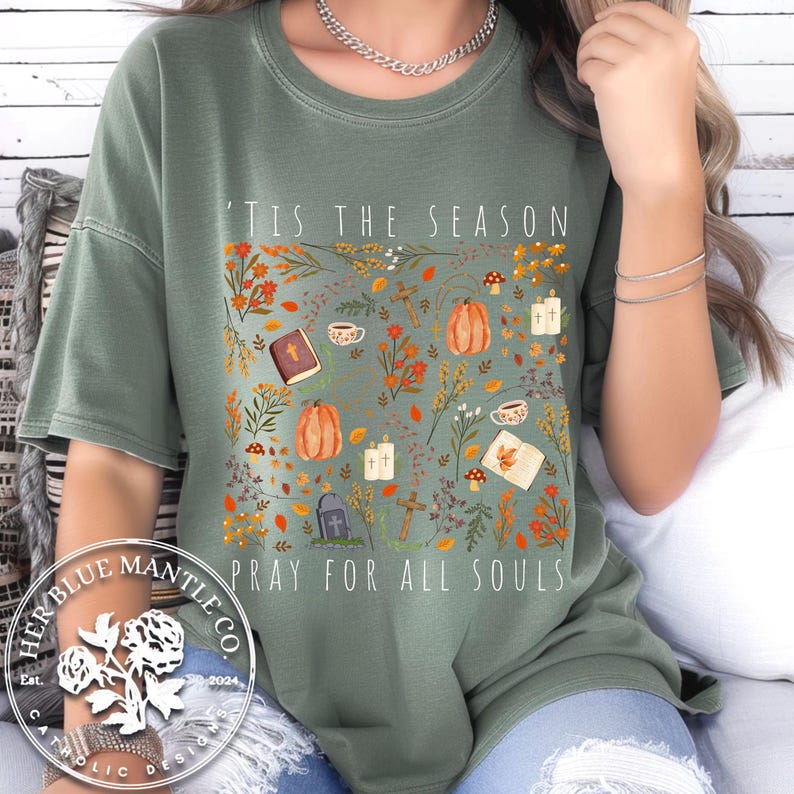 May include: A green t-shirt with a fall-themed design featuring a variety of autumnal elements, including leaves, pumpkins, and crosses. The text "'Tis the season" and "Pray for all souls" is printed on the shirt.