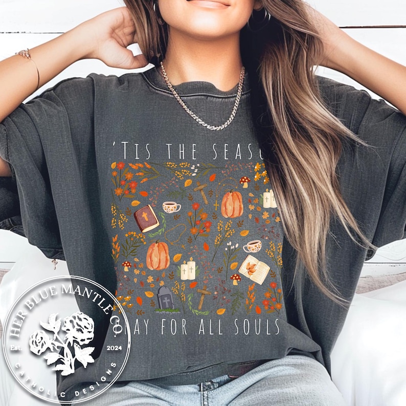 May include: A gray t-shirt with a fall-themed design featuring pumpkins, leaves, and a cross. The text on the shirt reads "'Tis the season to pray for all souls".