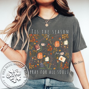 May include: A gray t-shirt with a fall-themed design featuring pumpkins, leaves, and crosses. The text "Tis the season" and "Pray for all souls" is printed on the shirt.