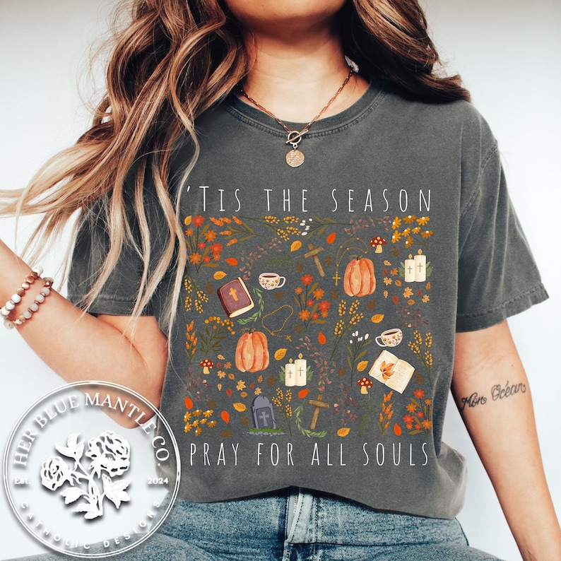 May include: A gray t-shirt with a fall-themed design featuring pumpkins, leaves, and crosses. The text "Tis the season" and "Pray for all souls" is printed on the shirt.
