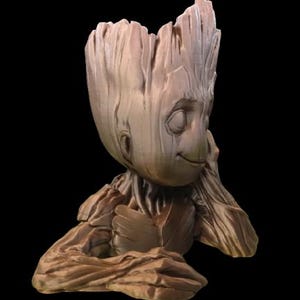 Puede incluir: Una figurita de madera marrón de un personaje de la película Guardianes de la Galaxia. El personaje está sentado con la cabeza apoyada en la mano, con una expresión pensativa. La figurita es una maceta, con un espacio hueco en la parte superior de la cabeza.