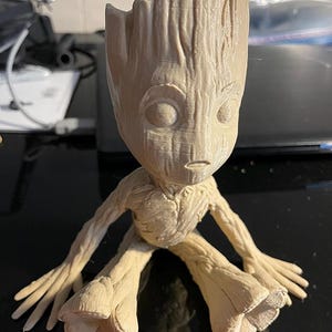 Puede incluir: Una figurita impresa en 3D de color beige de un personaje de la franquicia de películas Guardianes de la Galaxia. El personaje está sentado con las piernas cruzadas y tiene una superficie detallada y texturizada.
