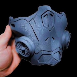 Może przedstawiać: Maseczka do twarzy w kolorze ciemnoniebieskim, wydrukowana w technologii 3D, o futurystycznym designie. Maseczka ma duży, geometryczny przód z małym wentylatorem z boku.
