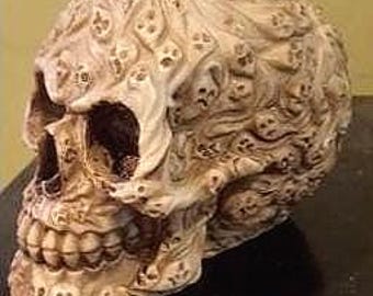 Könnte beinhalten: Eine detaillierte, cremefarbene Totenkopfskulptur mit strukturierter Oberfläche. Der Schädel ist mit kleinen, sich wiederholenden Totenkopfmustern bedeckt. Die Skulptur steht vor einem dunklen Untergrund und einem hellgelben Hintergrund.
