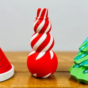 Pode incluir: Três decorações de Natal: um boneco de neve listrado de vermelho e branco, uma árvore de Natal verde e um chapéu de Papai Noel listrado de vermelho e branco.