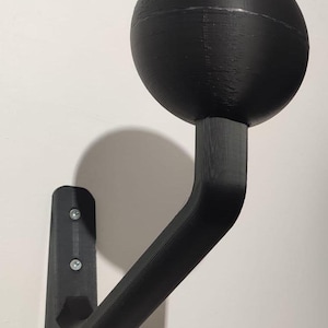 Puede incluir: Un gancho de pared negro con una bola redonda en la parte superior. El gancho está hecho de plástico y tiene un acabado liso y mate.