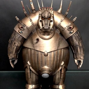 Puede incluir: Una figura de robot de color bronce con un cuerpo grande y redondo y una cabeza detallada. El robot tiene una corona con púas y múltiples brazos con diseños intrincados.