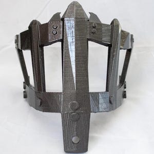 May include: A dark gray, 3D-printed crown-like headpiece. The design features a central vertical element and arched supports, with rivet-like details. The headpiece has a textured surface and a geometric aesthetic.