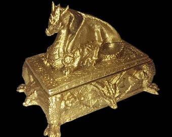 May include: A gold-colored dragon figurine sits atop a gold-colored box with intricate details. The box has four feet that resemble dragon claws.