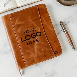May include: A tan leather notebook cover featuring a vertical stitched panel design. The front is embossed with the text "YOUR LOGO HERE" in a bold, dark font, and the initials "CVB" are embossed in the bottom right corner of the cover.
