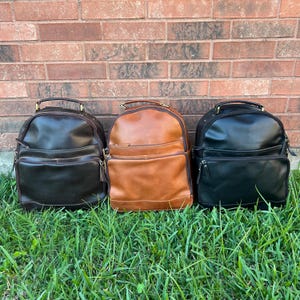 May include: Three leather backpacks in different colors: black, brown, and dark brown. The backpacks have multiple pockets and zippers.