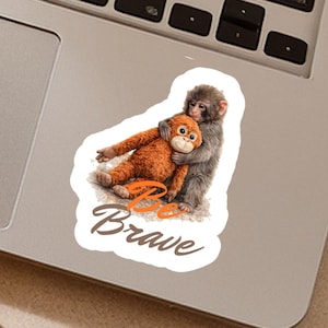 May include: A sticker featuring a gray monkey hugging an orange stuffed animal. The sticker has the words "Be Brave" in a handwritten style font. The sticker is on a silver laptop.