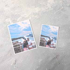 May include: Two rectangular photo prints featuring a family posing in front of a waterfall under a blue sky. The prints have white borders and are placed on a textured gray surface. The family is wearing casual clothing.