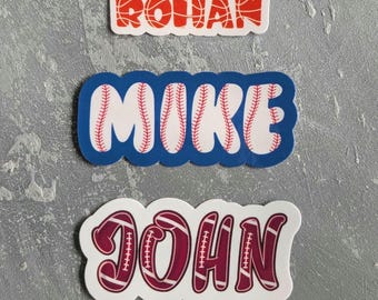 May include: Three name stickers: "ROHAN" in orange basketball design, "MIKE" in baseball design with a blue border, and "JOHN" in a maroon football design.