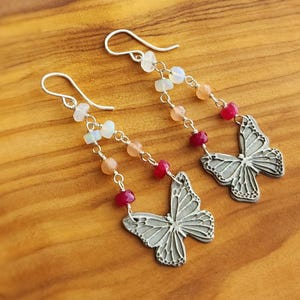 May include: A pair of dangle earrings featuring silver-toned metal butterfly charms suspended from curved ear wires. Each earring includes a delicate chain adorned with small, multi-colored gemstone beads in shades of pink, peach, and translucent white.