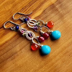 May include: Silver dangle earrings with a variety of colorful gemstones. The earrings feature blue, red, orange, purple, and green stones, with a large turquoise bead at the bottom of each earring. The earrings are set against a wooden background.