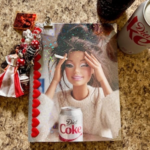 May include: A rectangular notebook featuring a holographic cover image of a doll with messy hair, wearing a white sweater, holding a cigarette, and sitting behind a Diet Coke can. The spine is decorated with red heart charms and a doll figurine.