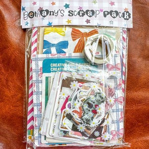 May include: A clear plastic bag filled with scrapbooking supplies. The bag contains patterned paper, stickers, and washi tape. The top of the bag has a decorative header that reads "bethany's scrap pack" in a whimsical font, with star and heart accents.
