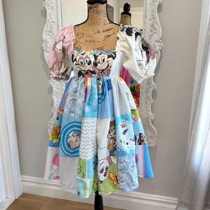 May include: A patchwork dress featuring Disney characters, displayed on a mannequin. The dress has a square neckline, puffed sleeves, and a skirt composed of various fabric squares with cartoon images. The color palette includes pink, blue, and white.