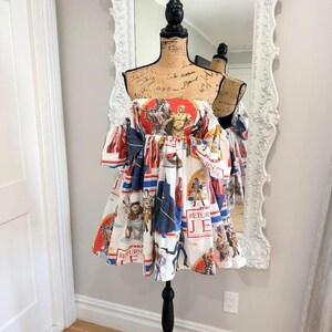 May include: An off-the-shoulder dress featuring a white base with a colourful Star Wars Return of the Jedi print. The fabric displays various characters and the text Return of the Jedi in red and blue accents. The dress has short puff sleeves.