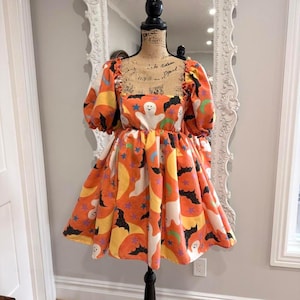 May include: A short-sleeved dress featuring an orange background with a repeating pattern of white ghosts, black bats, yellow crescent moons, and purple stars. The garment has a square neckline and puffed sleeves, displayed on a mannequin.