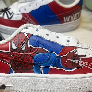 May include: Custom-painted white sneakers featuring a Spider-Man design. The shoes have red, blue, and white accents, with a detailed illustration of Spider-Man on the side. The name "WYATT" is written on the back of one shoe.
