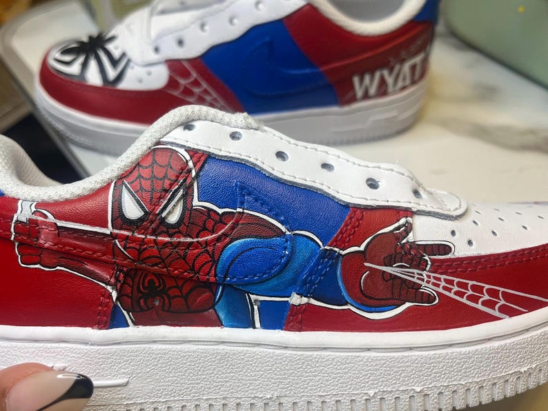 May include: Custom-painted white sneakers featuring a Spider-Man design. The shoes have red, blue, and white accents, with a detailed illustration of Spider-Man on the side. The name "WYATT" is written on the back of one shoe.