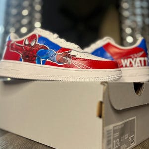 May include: White sneakers with red and blue accents, featuring a Spiderman graphic on the side. The shoes are on top of a gray shoe box. The name "WYATT" is printed on the back of one shoe. The shoes are size 13.