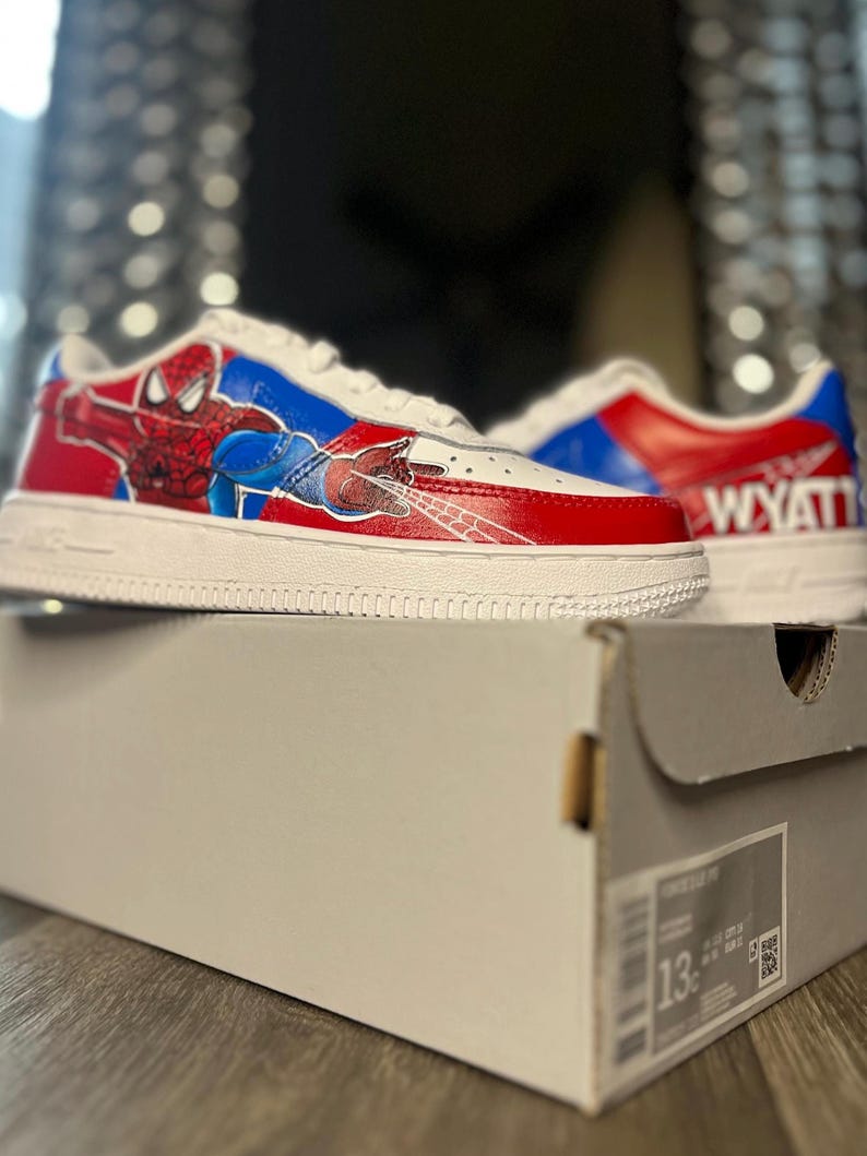 May include: White sneakers with red and blue accents, featuring a Spiderman graphic on the side. The shoes are on top of a gray shoe box. The name "WYATT" is printed on the back of one shoe. The shoes are size 13.
