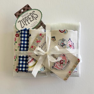 May include: A bundle of folded fabric squares featuring floral and gingham patterns, tied with a white ribbon. Included are a pink teapot-shaped charm, a Cath Kidston branded tag, and a Happy Zippers by Lori Holt packaging header card.