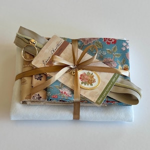Può includere: Un kit per il fai-da-te con tessuto piegato a motivi floreali blu, una cerniera beige e un foglio di feltro bianco. Un ciondolo Jane Austen è fissato su un cartoncino, il tutto legato con un nastro dorato. Testo sul cartoncino: Jane Austen Charm.