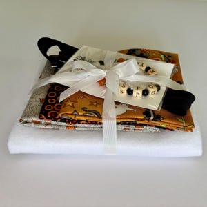 May include: A stack of folded fabric featuring orange, black, and white patterns with ghost and star motifs, topped with a white ribbon-tied package containing a beaded bracelet with wooden letter beads spelling SPOOKY and black beads.