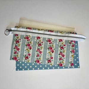 May include: A rectangular fabric pouch project featuring a white zipper with a silver ring pull. The pouch displays a floral pattern of pink roses and yellow flowers on vertical light blue stripes, layered over a blue fabric with white polka dots.