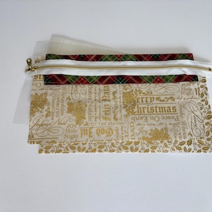 May include: A rectangular pouch featuring a white zipper with a gold pull. The top section has a red, green, and gold plaid pattern. The bottom section is cream-colored fabric with gold text including Merry Christmas and various holiday phrases.
