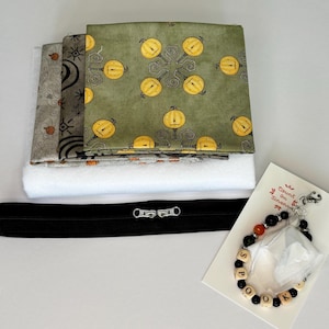 May include: A craft kit containing folded fabric squares with yellow pumpkin and star patterns, a white batting sheet, a black zipper with two silver pulls, and a beaded charm on a card labeled Count on Linen with the word SPOOK spelled out in beads.