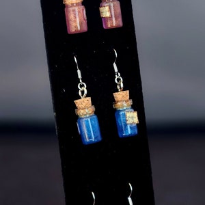 May include: Three pairs of dangle earrings featuring small glass bottles with cork stoppers. The bottles are filled with pink, blue, and red glitter.