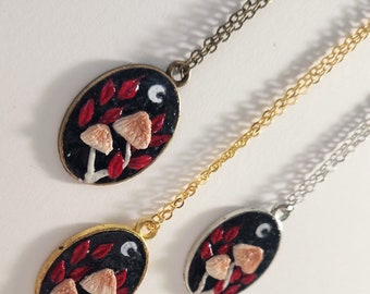 May include: Three silver, gold, and bronze necklaces with oval pendants featuring a black background with red leaves, white mushrooms, and a white crescent moon.