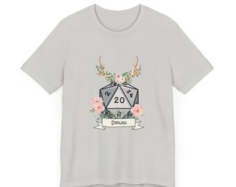 Puede incluir: Una camiseta gris claro con un gráfico de un dado de 20 caras con cuernos y flores. El dado tiene los números 2, 8, 14 y 20. El texto "DRUID" está en una pancarta debajo del dado.