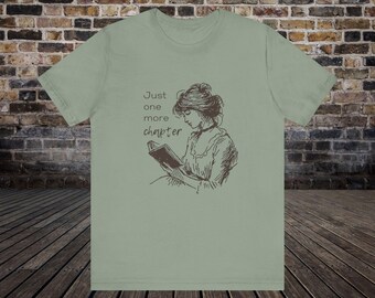 May include: A light green t-shirt featuring a brown illustration of a woman reading a book. The text "Just one more chapter" is printed on the shirt.