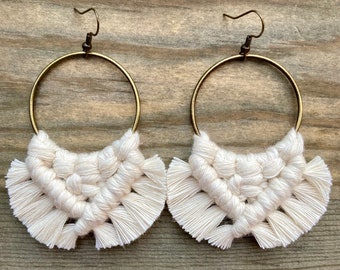 May include: A pair of handmade macrame earrings. Each earring features a cream-colored, woven macrame design suspended from a gold-colored hoop. The macrame has a fan-like shape with a fringe at the bottom. The earrings are displayed on a wooden surface.