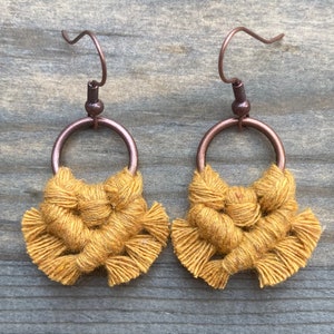 May include: A pair of handmade macrame earrings. Each earring features a copper-colored hoop with a woven, mustard-yellow macrame design. The earrings have copper-colored ear wires and are displayed on a wooden surface.