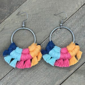 May include: A pair of handmade macrame hoop earrings. Each earring features a silver-colored hoop with a colorful fringe of blue, light blue, pink, and orange yarn. The earrings are displayed on a wooden surface.