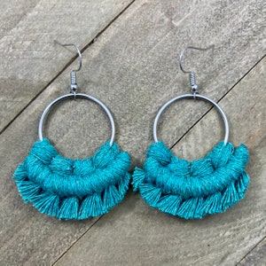 May include: A pair of turquoise macrame earrings with silver-colored hoops. The earrings feature a half-circle of knotted turquoise thread, creating a fringe effect. The earrings are designed for pierced ears.