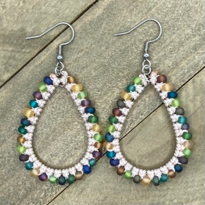 May include: A pair of teardrop-shaped earrings. Each earring features a white macrame design, adorned with rows of colorful beads in shades of blue, green, yellow, and purple. The earrings have silver-colored ear wires.