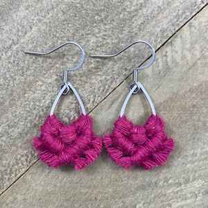 May include: A pair of handmade magenta macrame earrings. Each earring features a silver teardrop-shaped frame with a woven, textured design. The earrings have silver-colored ear wires for pierced ears. The earrings are displayed on a wooden surface.